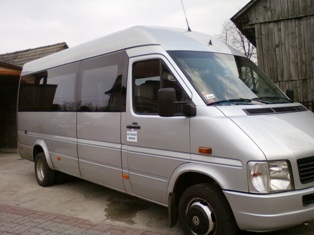 Srebrny bus z przyciemnianymi szybami, zaparkowany przed drewnianym budynkiem. Na karoserii widoczna naklejka z informacją o wynajmie.