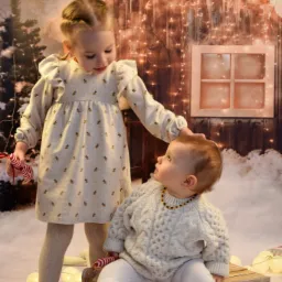 Dwie dziewczynki w świątecznej scenerii: starsza w sukience z motywem ananasów trzyma laskę cukrową i dotyka głowy młodszego dziecka siedzącego na drewnianych sankach, w tle oświetlona chatka...