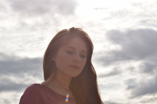 Portret młodej kobiety z długimi, ciemnymi włosami na tle pochmurnego nieba, delikatne światło słoneczne w prawym dolnym rogu.