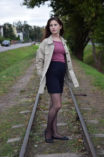 Portret kobiety w beżowym płaszczu i czarnych rajstopach stojącej na starych torach kolejowych, z zielenią w tle