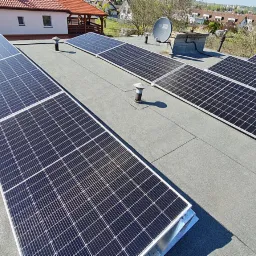 Panele fotowoltaiczne zamontowane na płaskim dachu pokrytym papą, widoczne kominki wentylacyjne i antena satelitarna, w tle zabudowa jednorodzinna.