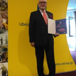 Mężczyzna w garniturze prezentuje certyfikat 'Million Dollar Round Table AVIVA Uczestnik 2019' na tle żółtego baneru z logo Aviva i napisem 'Ubez'.