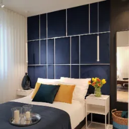 Nowoczesna sypialnia z granatowym panelem ściennym obitym welurem, białymi lampami wiszącymi, pościelą z kolorowymi poduszkami i bukietem tulipanów na stoliku nocnym.