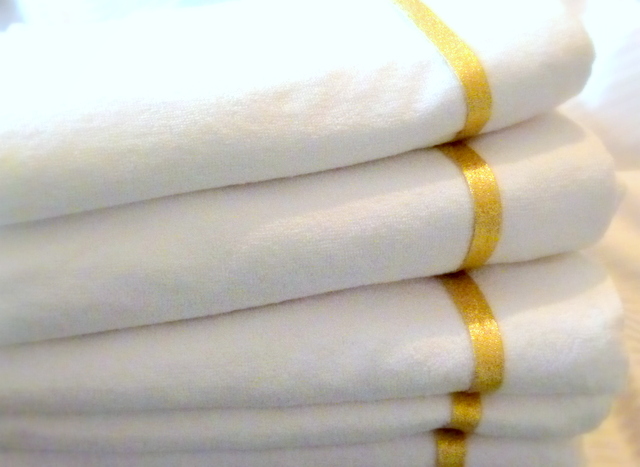 Stos białych, miękkich ręczników frotte z dekoracyjnym złotym paskiem na brzegach, ułożonych równo jeden na drugim.
