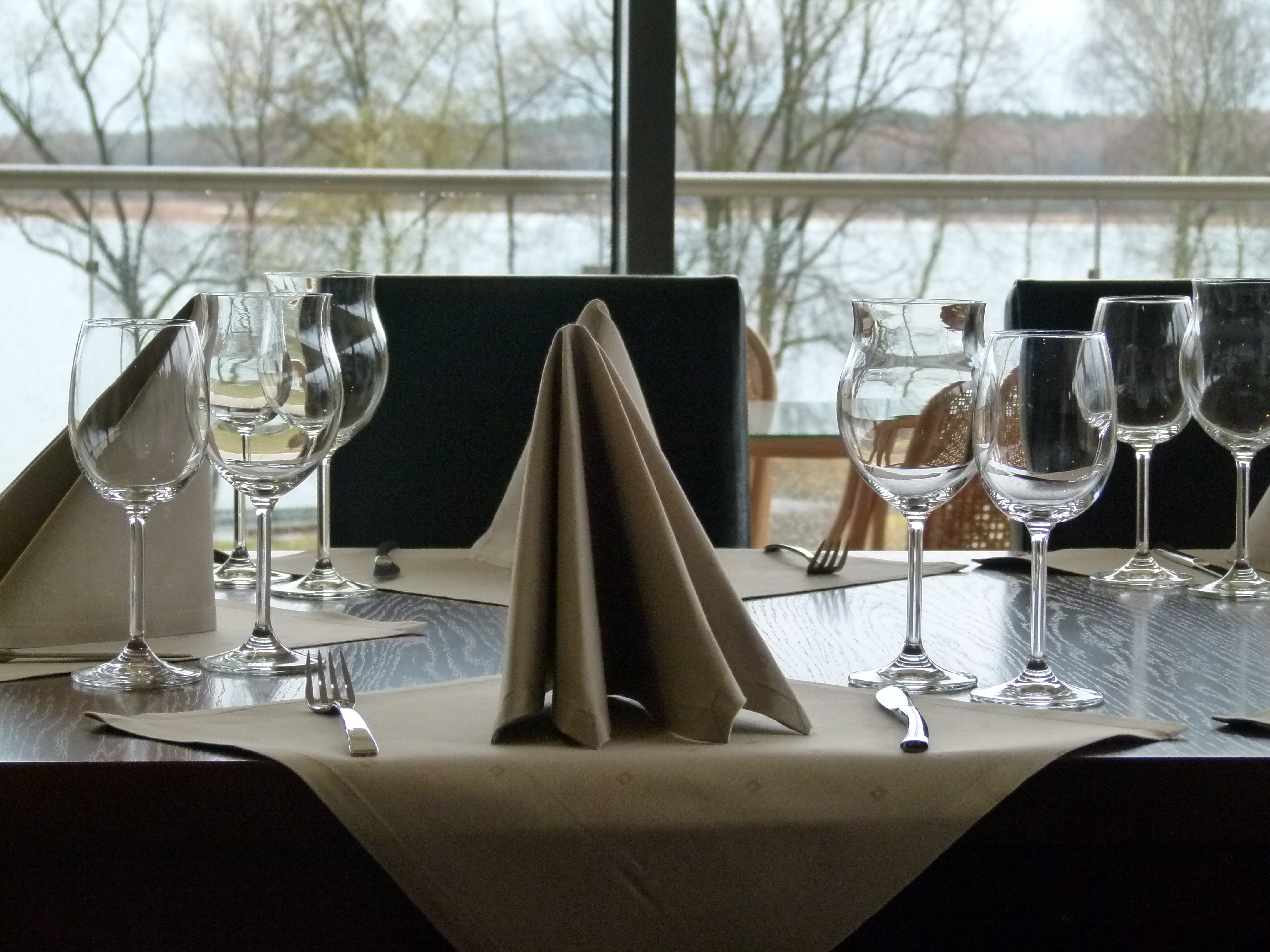 Nakryty stół w restauracji z kieliszkami, sztućcami i złożonymi serwetkami, widok na jezioro za oknem.