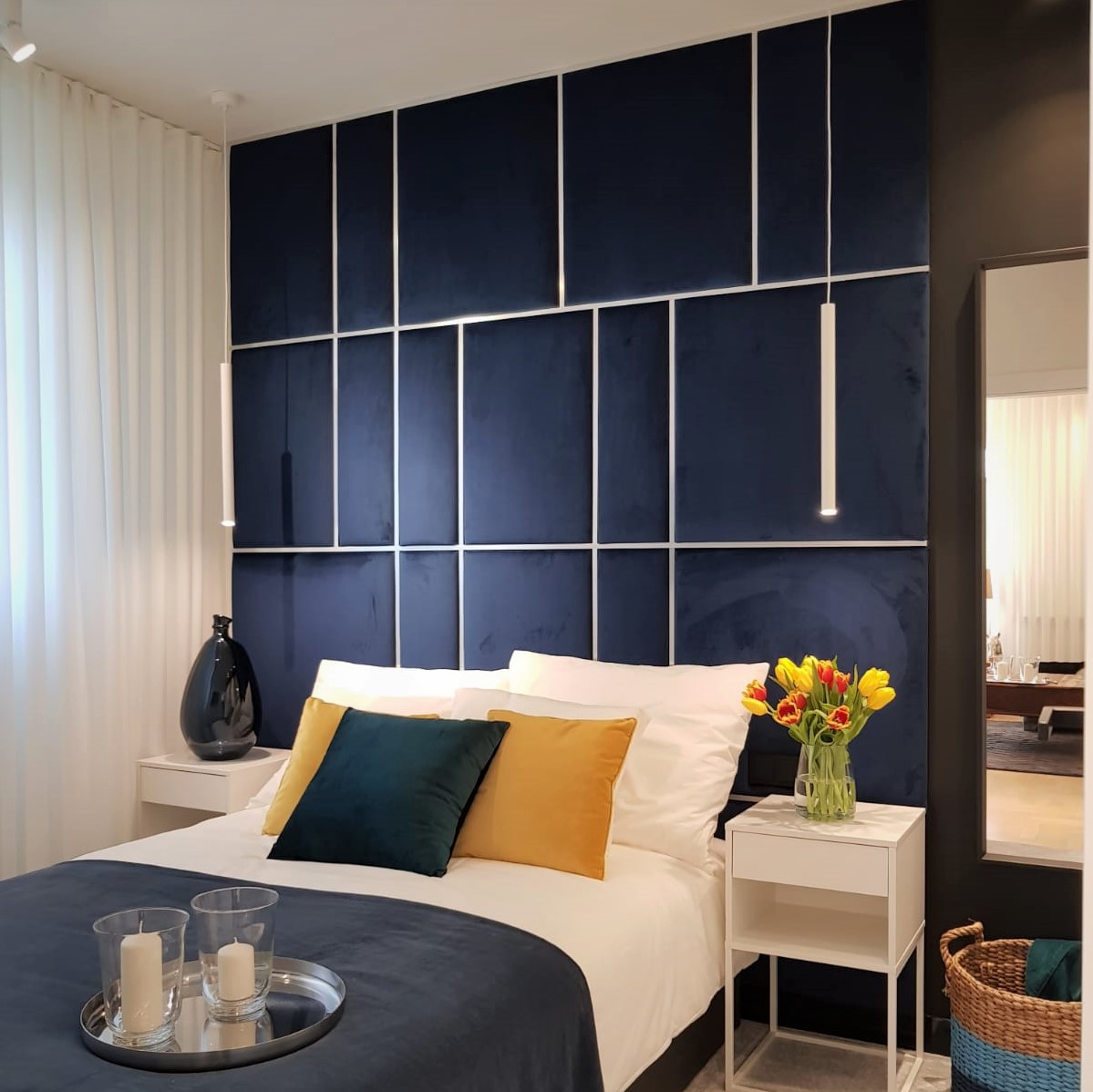 Nowoczesna sypialnia z granatowym panelem ściennym obitym welurem, białymi lampami wiszącymi, pościelą z kolorowymi poduszkami i bukietem tulipanów na stoliku nocnym.