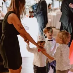 Kobieta z tatuażem lisa na ramieniu tańczy z dziećmi na przyjęciu, jedno z dzieci ma papierową koronę i czerwoną muszkę.