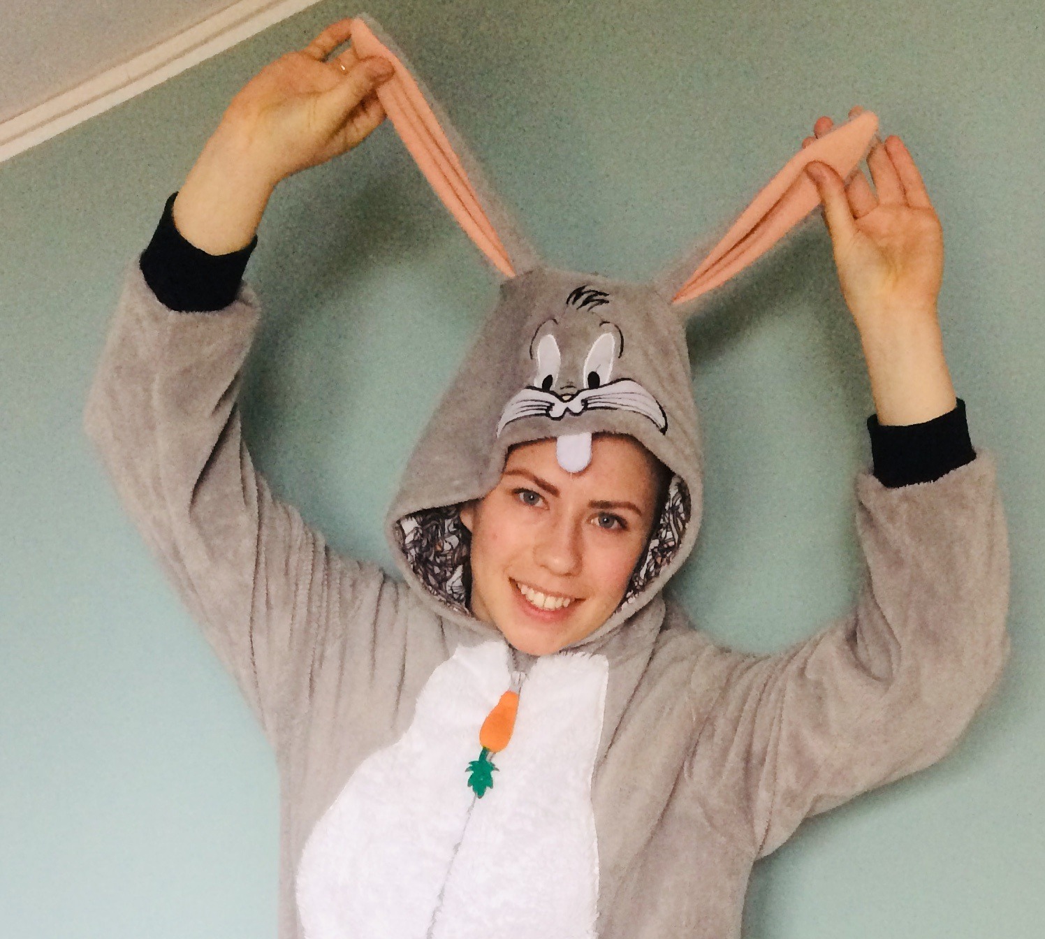 Uśmiechnięta kobieta w kostiumie pluszowego królika Bugs Bunny, z rękami uniesionymi do góry, pozuje na tle jasnoniebieskiej ściany.