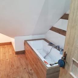 Remonty i dachy - Wnętrze łazienki w trakcie remontu: obudowana wanna płytkami imitującymi drewno, białe ściany ze skośnym sufitem, narzędzia glazurnicze na wannie, podłoga wyłożona panelami imitującymi drewno...