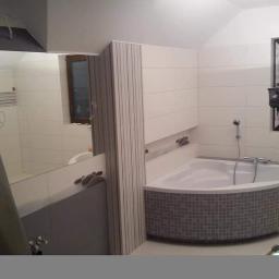 Remonty i dachy - Narożna wanna obłożona szarą mozaiką w łazience z białymi płytkami, widoczne narzędzia glazurnicze i odbicie w lustrze fragmentu łazienki.