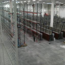 Remonty i dachy - Montaż regałów magazynowych w hali, widok z góry, częściowo zamontowane konstrukcje z czerwonymi elementami, pracownik w oddali, surowy beton na podłodze.