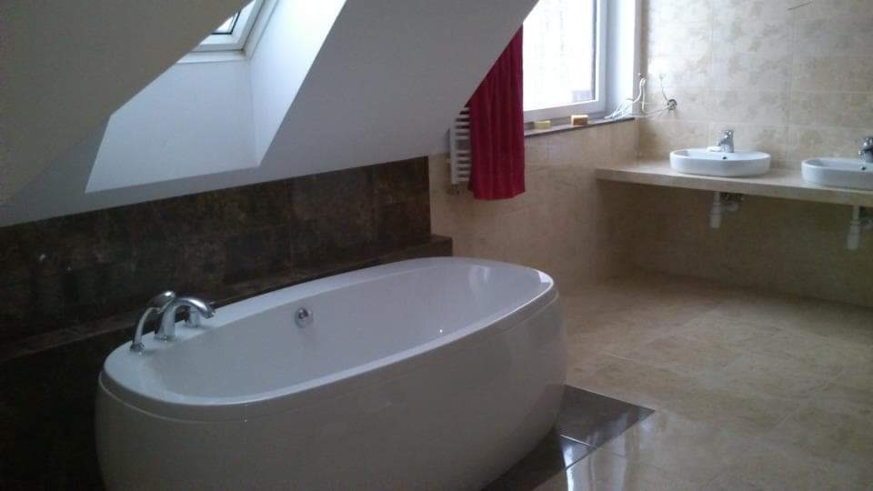 Nowoczesna łazienka na poddaszu z owalną wanną, jasnymi płytkami i dwoma umywalkami nablatowymi, oświetlona przez okno dachowe.