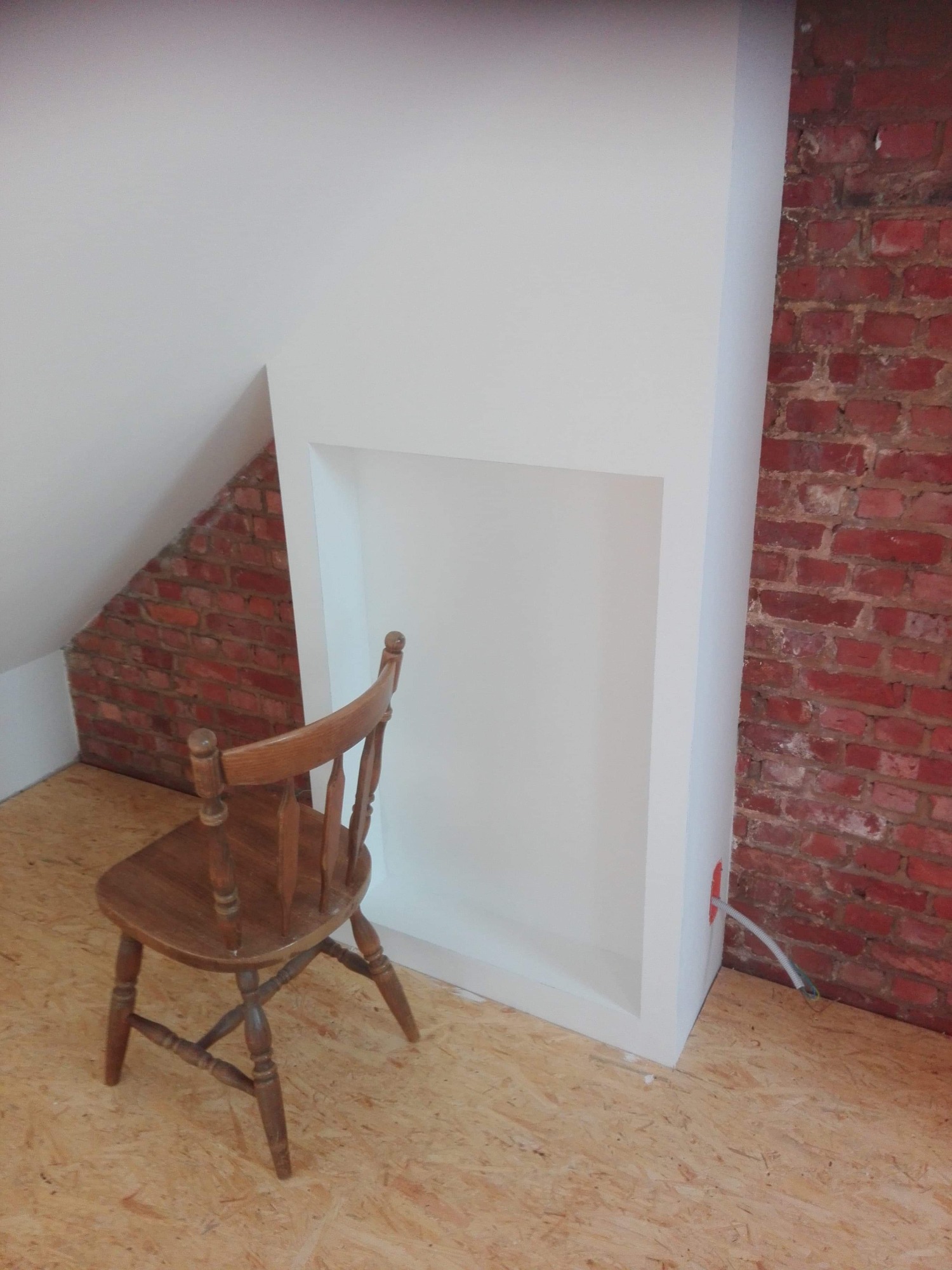 Poddasze z fragmentem ściany z czerwonej cegły, białą zabudową wnęki i drewnianym krzesłem na podłodze z płyt OSB, widoczny przewód elektryczny.