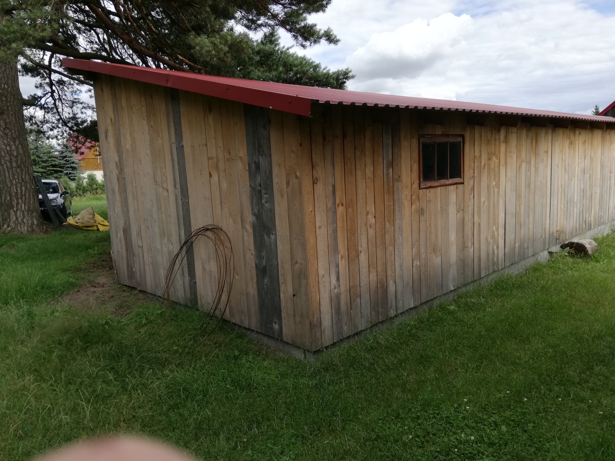 Długi, drewniany budynek gospodarczy z czerwonym dachem z blachy trapezowej, małe okno z ciemną szybą, otoczony zielonym trawnikiem, widoczne drzewo i fragment samochodu w tle.