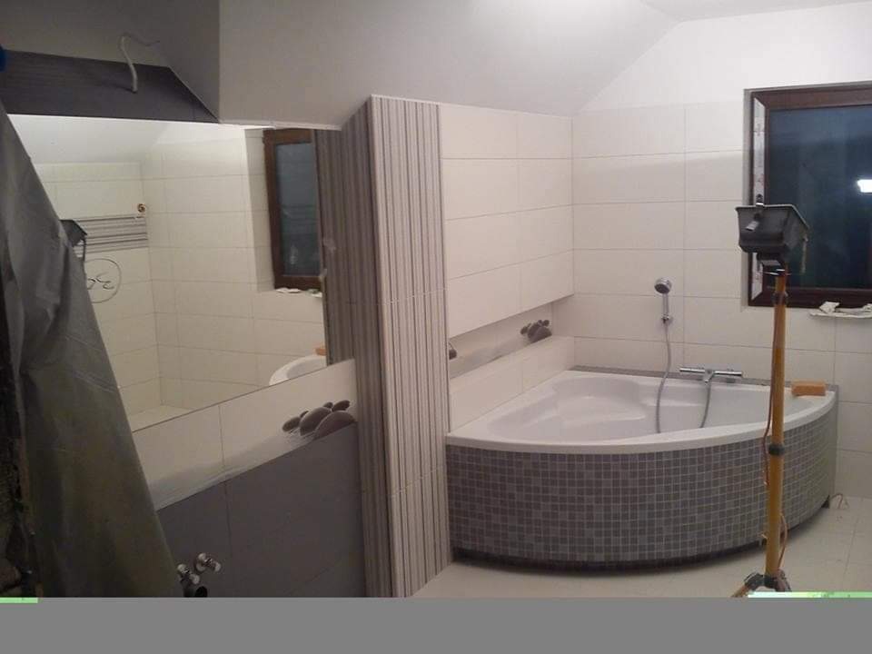 Narożna wanna obłożona szarą mozaiką w łazience z białymi płytkami, widoczne narzędzia glazurnicze i odbicie w lustrze fragmentu łazienki.