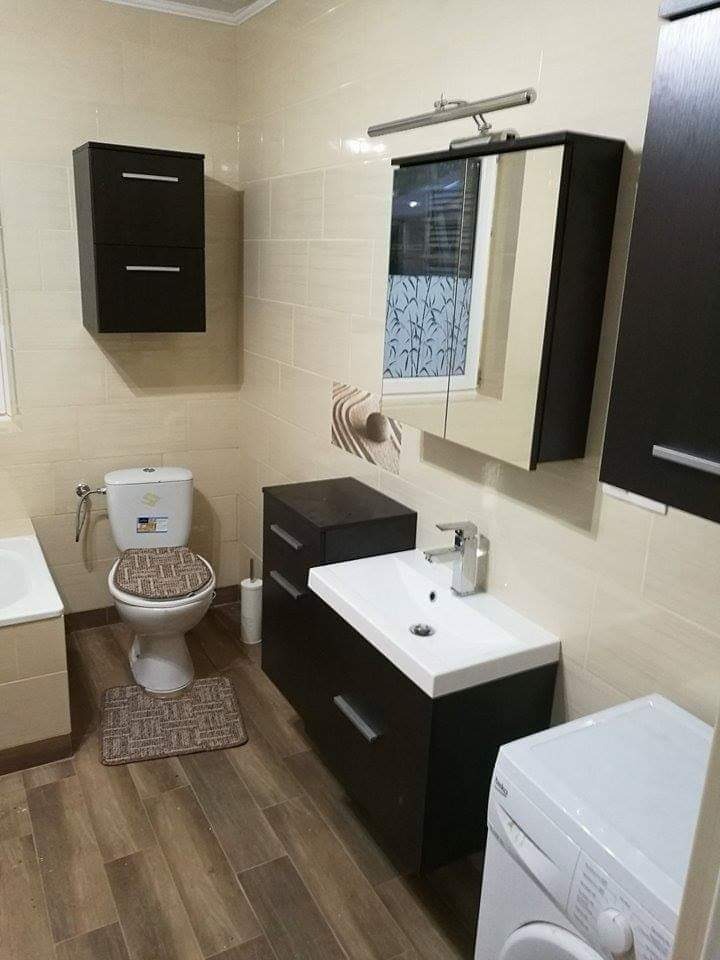 Umeblowana łazienka z brązowymi akcentami, obejmująca toaletę z dywanikiem, umywalkę z szafką, wiszącą szafkę i pralkę, wykończona jasnymi płytkami.