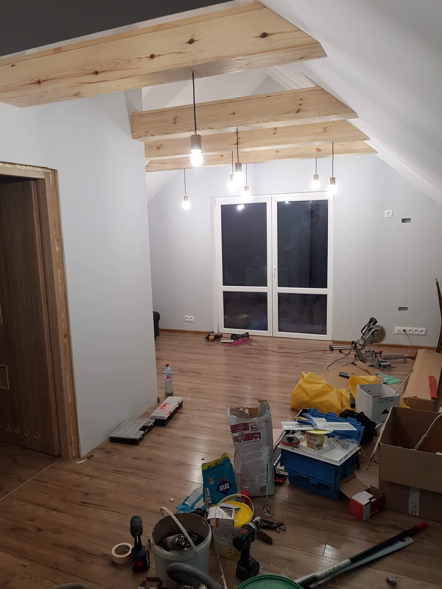 Wnętrze poddasza w trakcie prac wykończeniowych, widoczne drewniane belki stropowe z zamontowanym oświetleniem, okno balkonowe, częściowo pomalowane ściany oraz narzędzia i materiały budowlane...