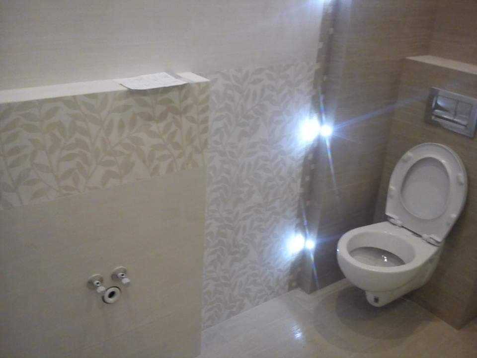 Nowoczesna toaleta wisząca z otwartą klapą, zamontowana na ścianie z beżowymi płytkami imitującymi drewno, obok ściana z wzorem liści, widoczne przyłącza wodne.