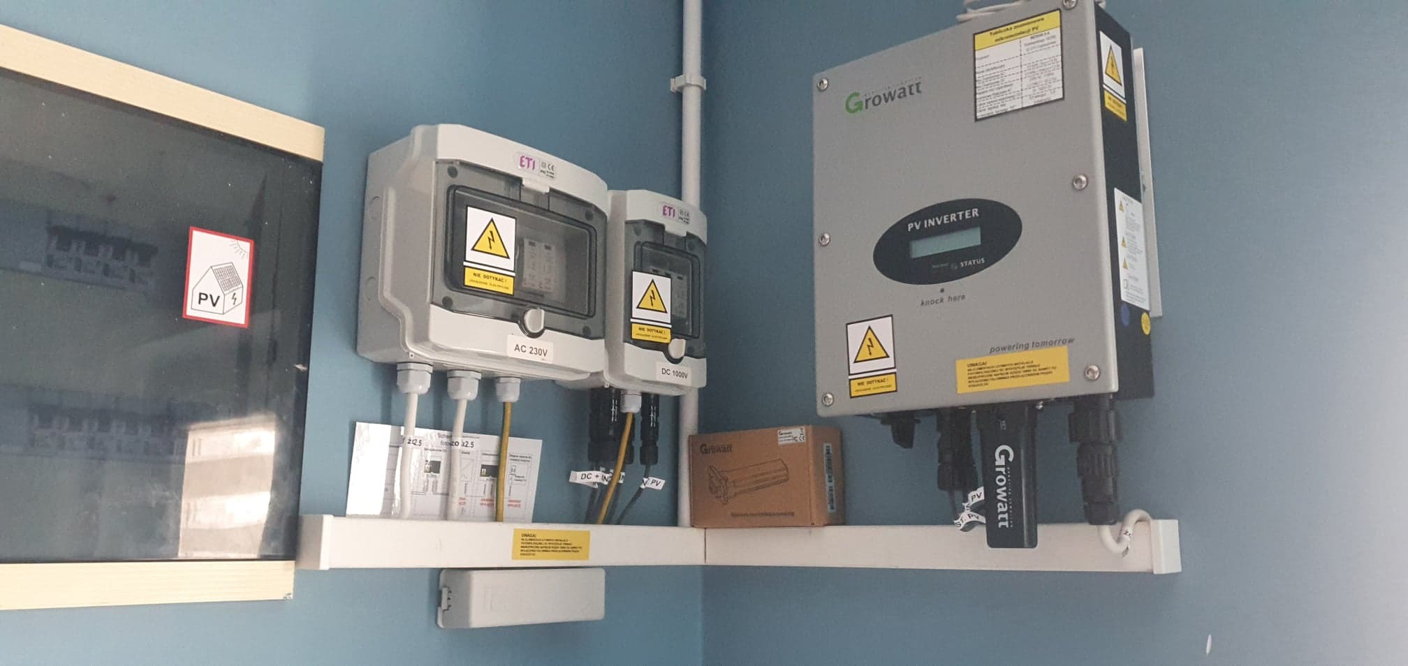 Instalacja fotowoltaiczna w pomieszczeniu: falownik Growatt, skrzynki ETI AC 230V i DC 1000V, zabezpieczenia, schematy elektryczne i naklejka PV na ścianie.