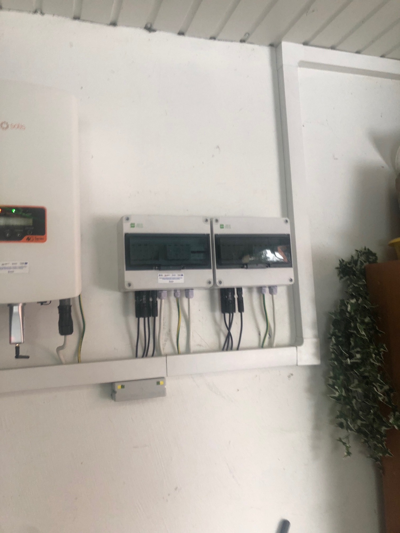 Biały inwerter Solis z podłączonymi kablami i dwa białe panele rozdzielcze zamontowane na białej ścianie wewnątrz budynku, widoczne elementy instalacji elektrycznej.