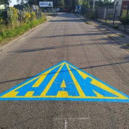 Niestandardowe oznakowanie poziome: duży, niebiesko-żółty trójkąt z napisem na asfalcie drogi dojazdowej.