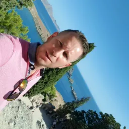 Mężczyzna w różowej koszulce polo z okularami przeciwsłonecznymi na tle błękitnego morza i zielonej roślinności, prawdopodobnie podczas remontu balkonu z widokiem.