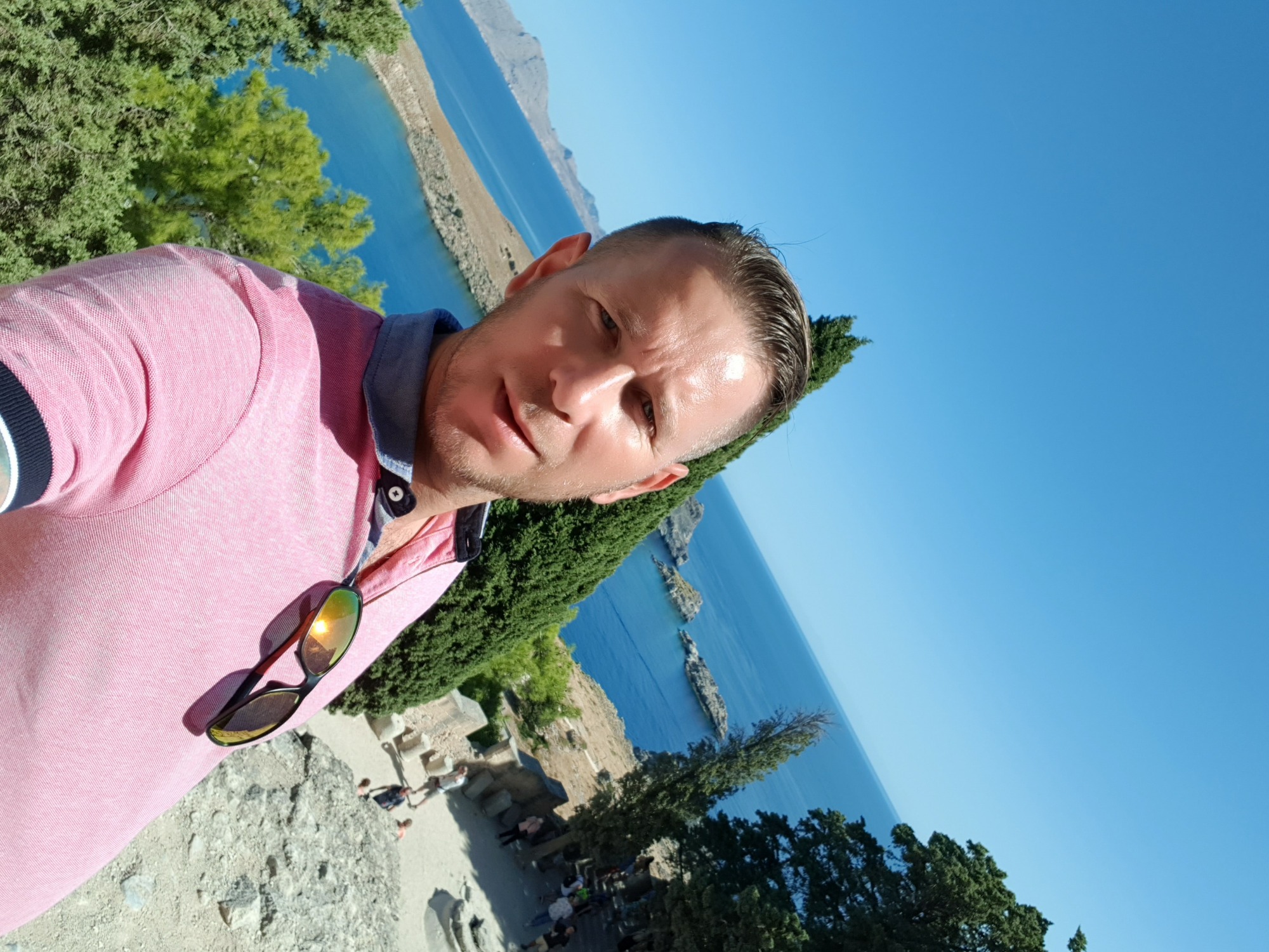 Mężczyzna w różowej koszulce polo z okularami przeciwsłonecznymi na tle błękitnego morza i zielonej roślinności, prawdopodobnie podczas remontu balkonu z widokiem.