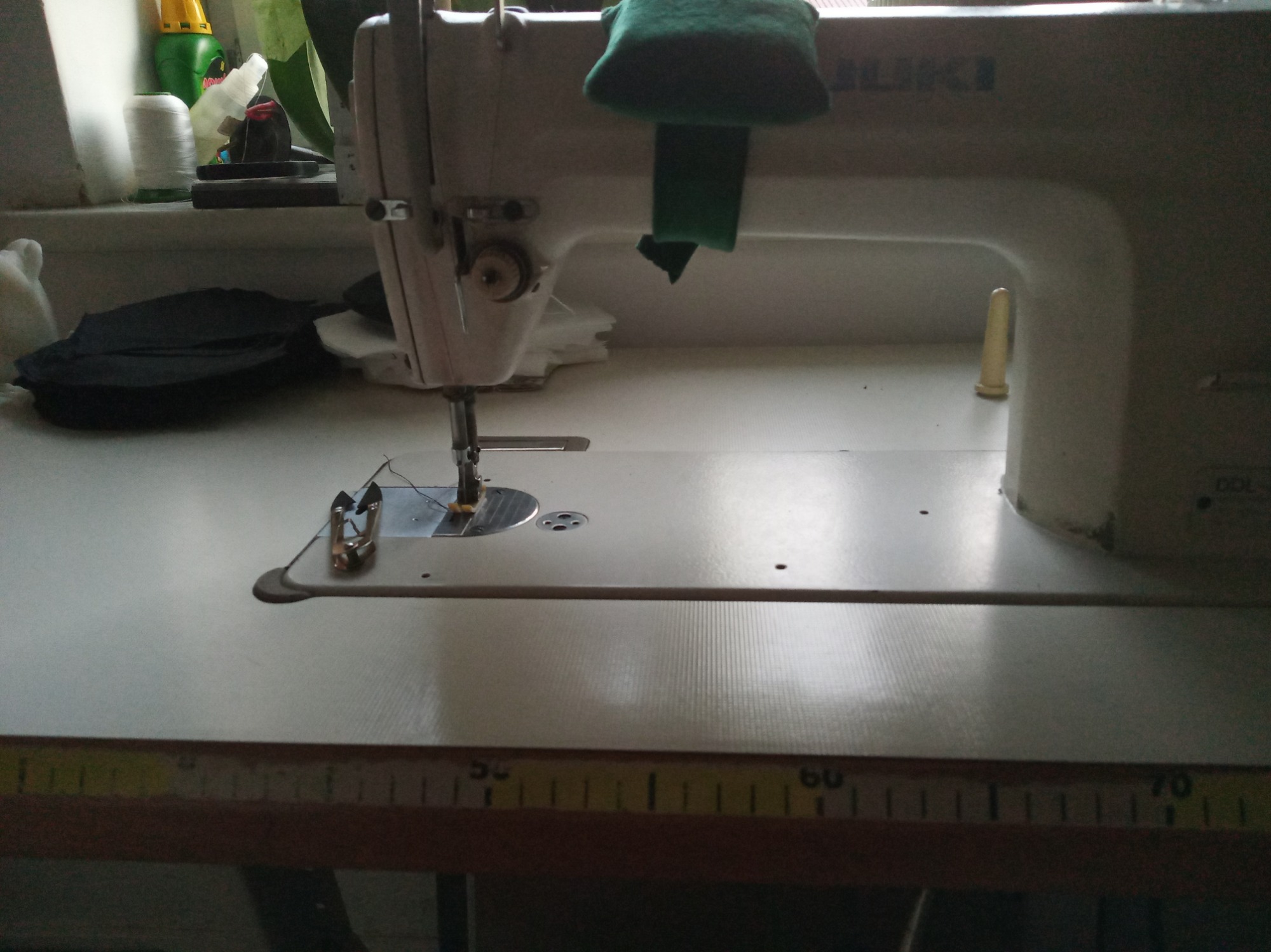 Bliski kadr na maszynę do szycia marki JUKI z zielonym materiałem na górze, widoczna igła w trakcie pracy i miarka na stole.