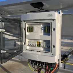 Rozdzielnica elektryczna Noark z otwartymi drzwiczkami, umieszczona pod panelami słonecznymi, z widocznymi bezpiecznikami i okablowaniem.