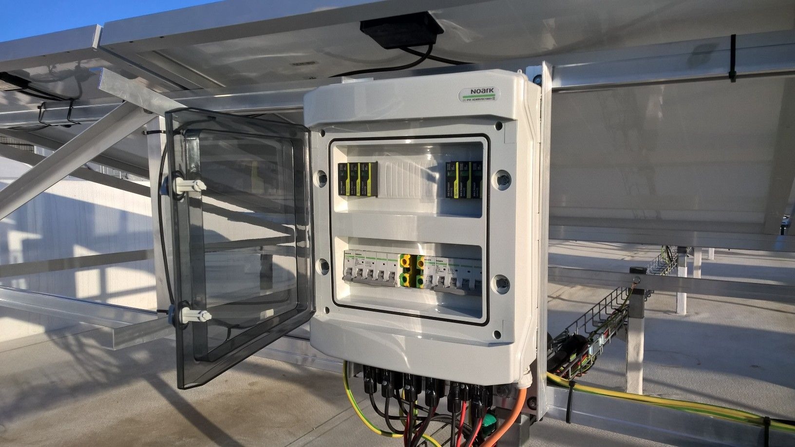 Rozdzielnica elektryczna Noark z otwartymi drzwiczkami, umieszczona pod panelami słonecznymi, z widocznymi bezpiecznikami i okablowaniem.