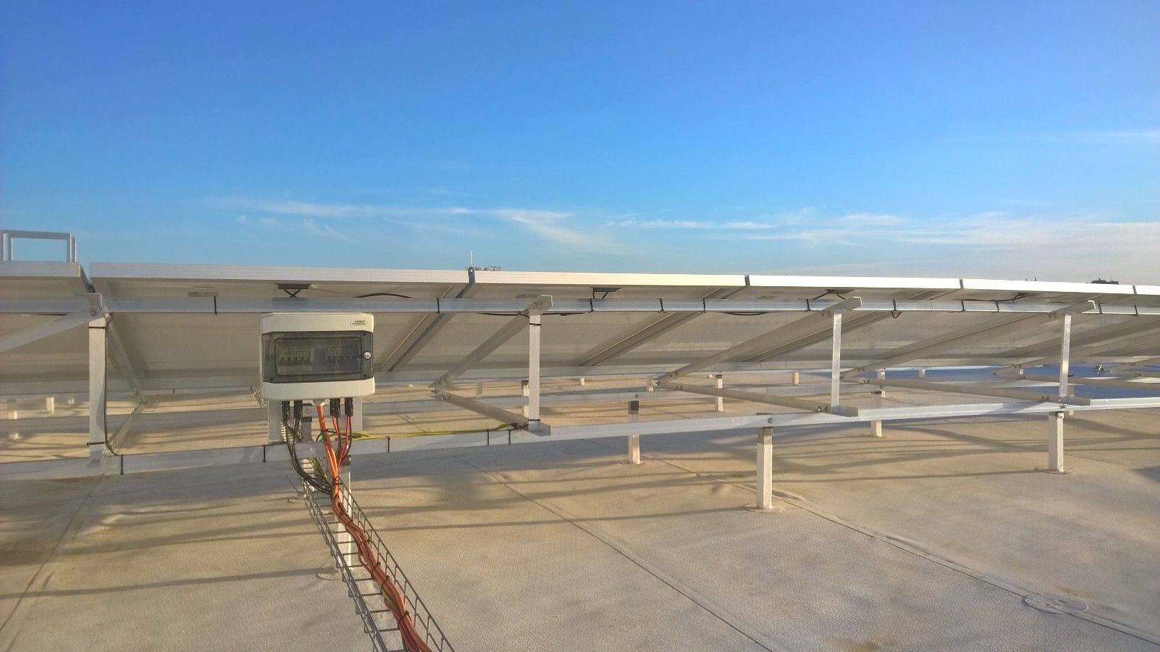 Montaż instalacji fotowoltaicznej: widok od spodu paneli, widoczna konstrukcja wsporcza, okablowanie w korytku i skrzynka elektryczna na dachu.