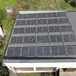 Widok z góry na instalację paneli słonecznych na płaskim dachu budynku z szarą papą, widoczna drabina oparta o ścianę budynku.