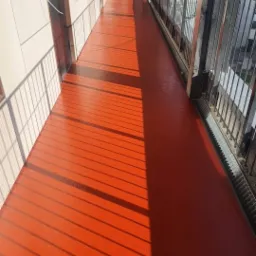 Pomarańczowa, gładka powierzchnia balkonu z żywicy, oświetlona promieniami słońca tworzącymi cienie kratownicy, z widocznymi metalowymi barierkami i fragmentem elewacji budynku.