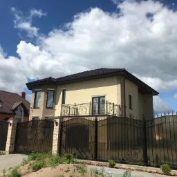 Dwukondygnacyjny dom z beżową elewacją, czarną balustradą balkonu i ozdobnym, kutym ogrodzeniem, widziany z poziomu gruntu w słoneczny dzień.