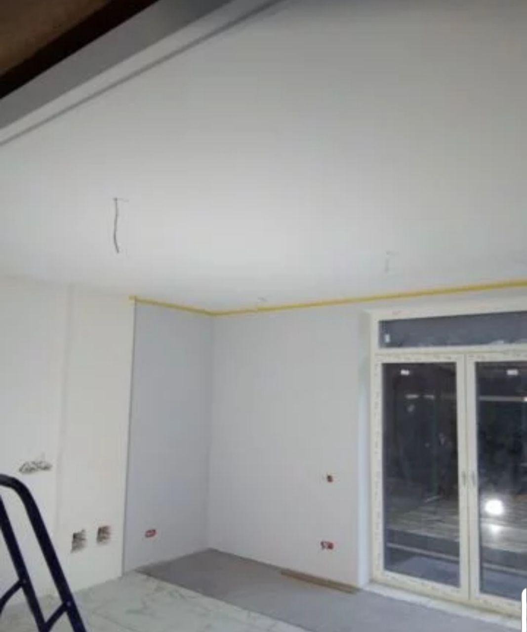 Pokój w trakcie prac wykończeniowych: ściany przygotowane do malowania, zabezpieczone taśmą malarską, widoczna drabina i okno balkonowe.