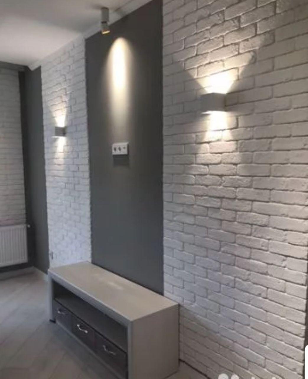 Ściana z białej cegły z zamontowanymi kinkietami, szara wnęka z oświetleniem punktowym i szafka RTV w minimalistycznym stylu.