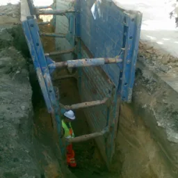 Głęboki wykop zabezpieczony niebieską obudową szalunkową; pracownik w kasku i kamizelce odblaskowej stoi wewnątrz wykopu, widoczny rowerzysta w tle.