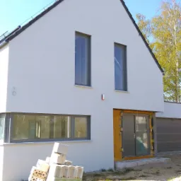 Nowoczesny dom jednorodzinny w trakcie budowy, widoczne szare okna, przygotowane materiały budowlane, wiosenne słońce.