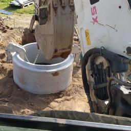 Kopex - Biała koparka umieszcza betonowy krąg w wykopie, widok z kabiny operatora, w tle zielone pole i drzewa.