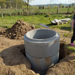 Betonowa studnia wkopana w ziemię, widoczny wykop, pryzma ziemi obok, w tle łąka i drzewa, osoba w różowych butach w prawym górnym rogu.