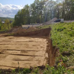 Przygotowany teren pod budowę domu jednorodzinnego, widoczne wytyczone granice fundamentów, w tle składowany piasek i częściowo wzniesione ściany fundamentowe.