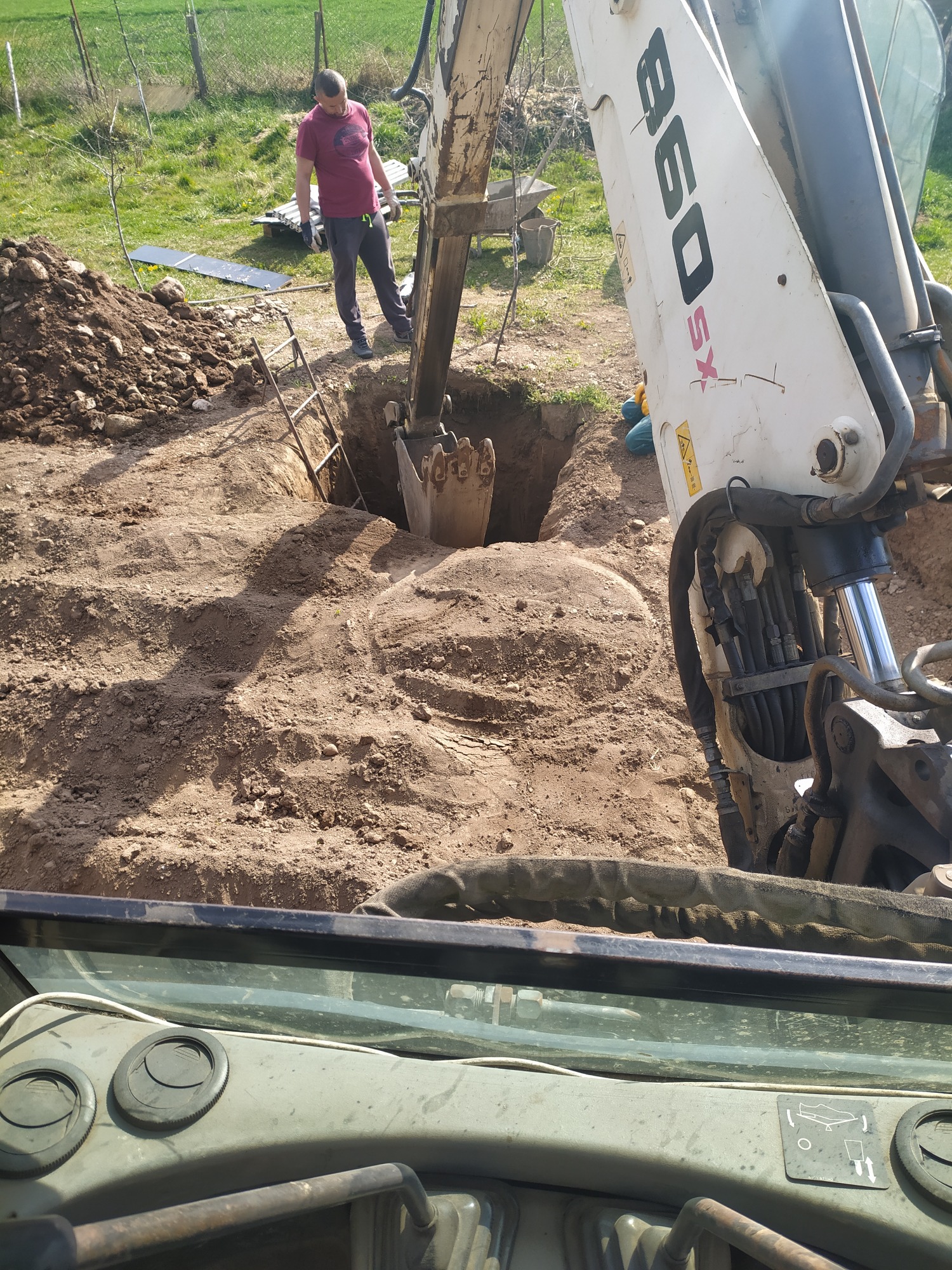 Koparka Liebherr 860 XS kopie głęboki dół w piaszczystej ziemi; w tle widoczna sterta ziemi, drabina i osoba nadzorująca prace.