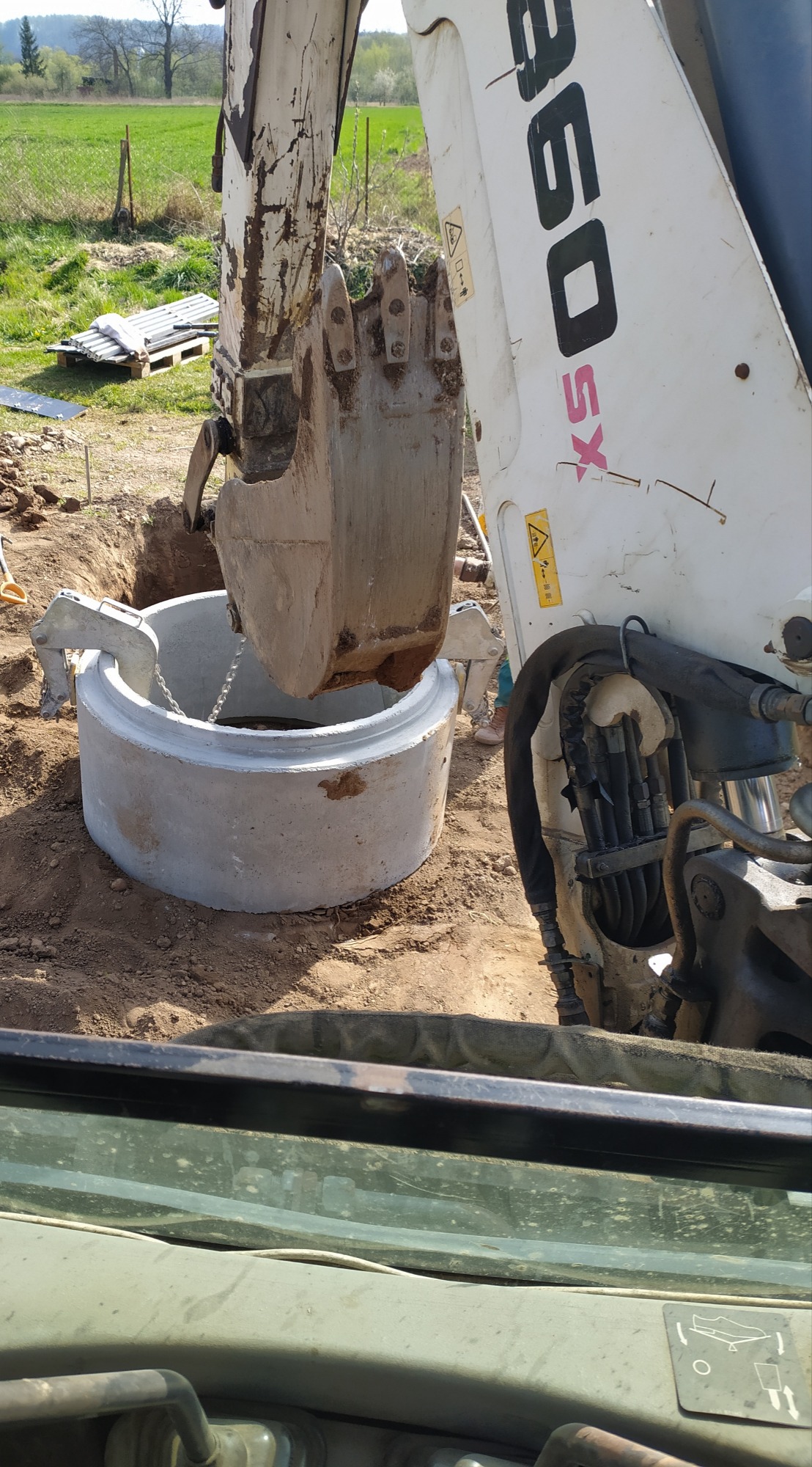 Biała koparka umieszcza betonowy krąg w wykopie, widok z kabiny operatora, w tle zielone pole i drzewa.