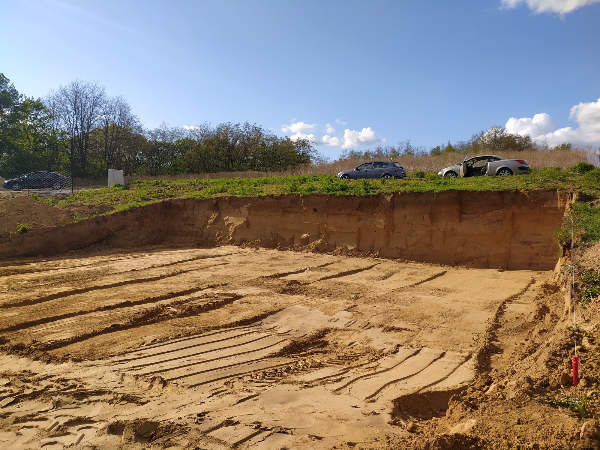 Wyrobisko ziemne z widocznymi śladami maszyn, na górze skarpy zaparkowane dwa samochody osobowe, błękitne niebo z chmurami w tle.