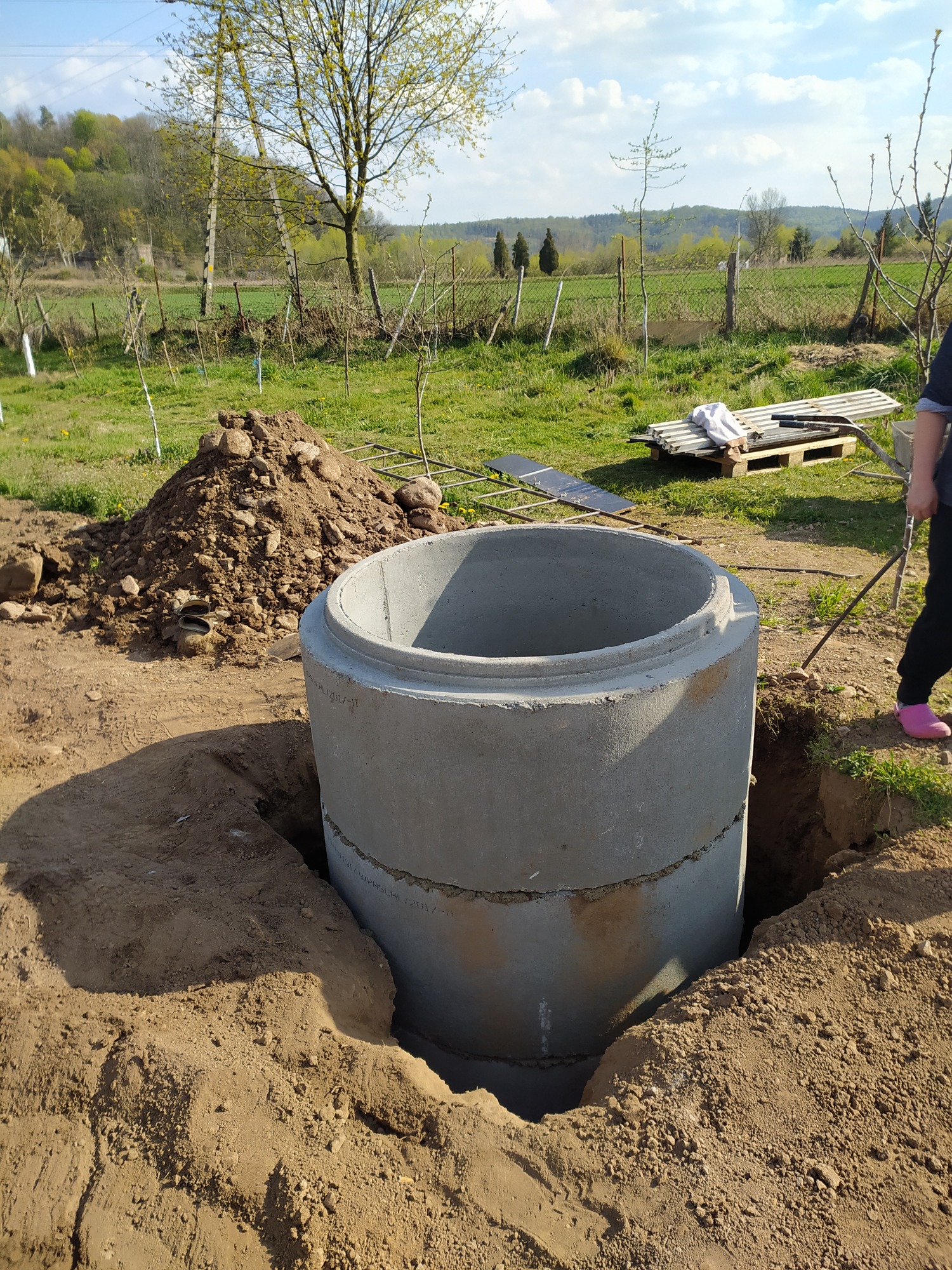 Betonowa studnia wkopana w ziemię, widoczny wykop, pryzma ziemi obok, w tle łąka i drzewa, osoba w różowych butach w prawym górnym rogu.
