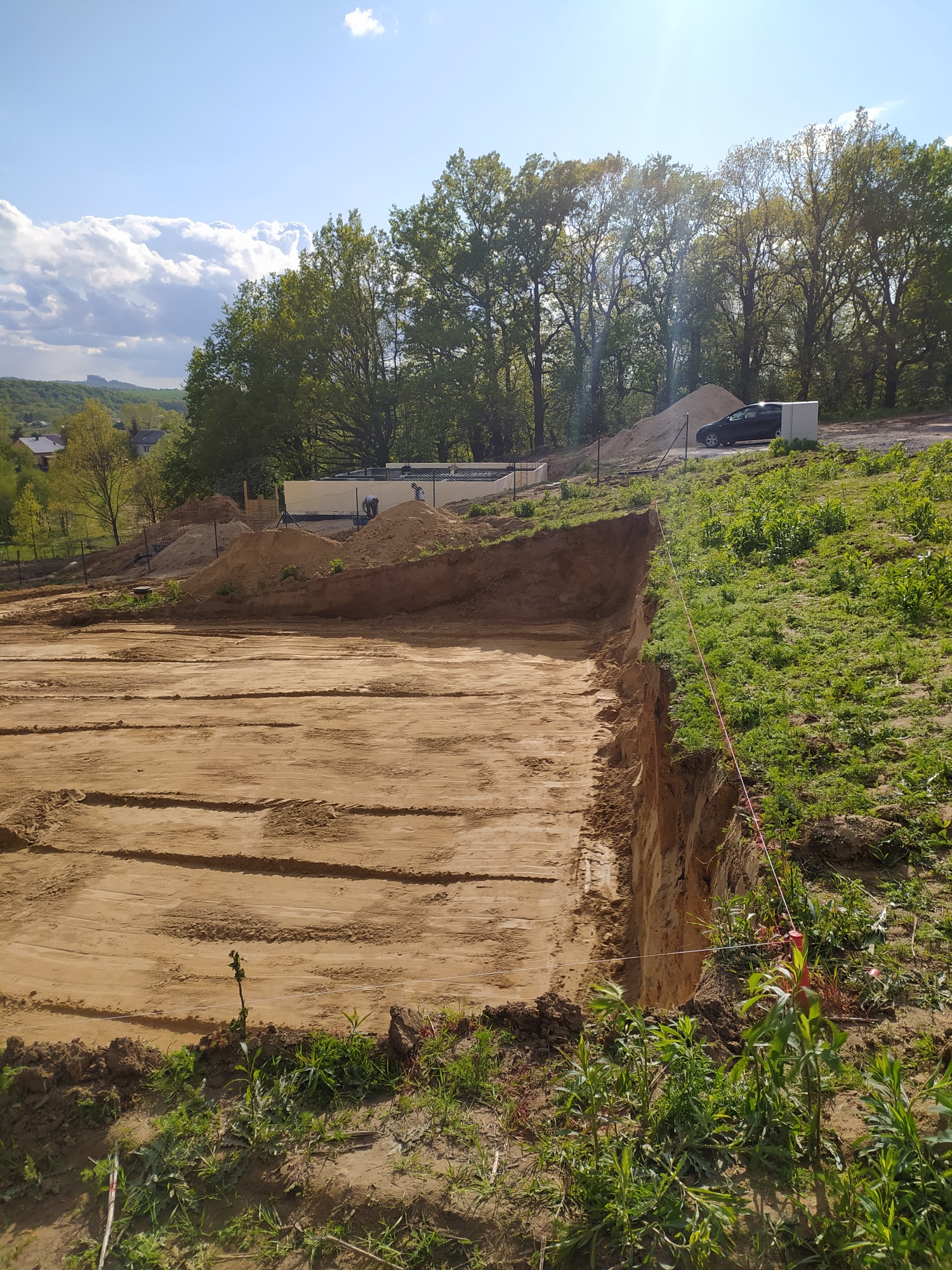 Przygotowany teren pod budowę domu jednorodzinnego, widoczne wytyczone granice fundamentów, w tle składowany piasek i częściowo wzniesione ściany fundamentowe.