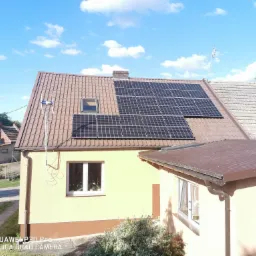 Panele słoneczne na brązowym dachu domu jednorodzinnego z anteną, widoczne okno dachowe i komin, błękitne niebo z chmurami w tle.