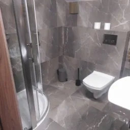 Wykończona łazienka z szarymi płytkami imitującymi marmur na ścianach i podłodze, kabina prysznicowa z zaokrąglonymi drzwiami, wisząca toaleta oraz kosz na śmieci w rogu.