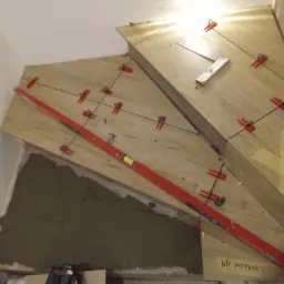 Montaż płytek na schodach z użyciem systemu poziomowania płytek (czerwone klipsy i kliny), poziomica w kolorze czerwonym, widoczna zaprawa murarska i kartka z napisem 'Nie dotykać!'