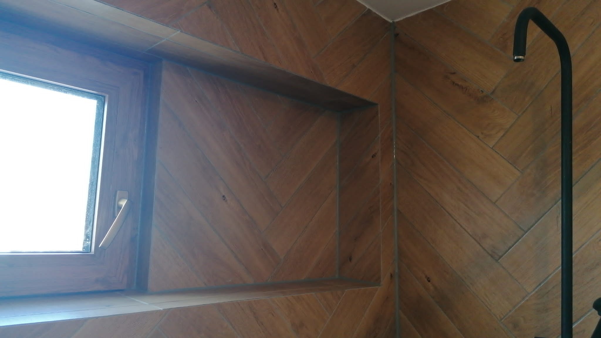 Łazienka wykończona płytkami imitującymi drewno ułożonymi w jodełkę, z oknem i czarną armaturą.